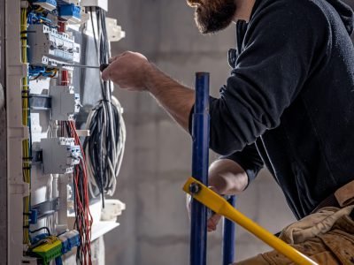 A male electrician works in a switchboard with an electrical connecting cable, connects the equipment with tools.